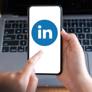 Person zeigt auf LinkedIn-Logo auf Smartphone mit Laptop im Hintergrund